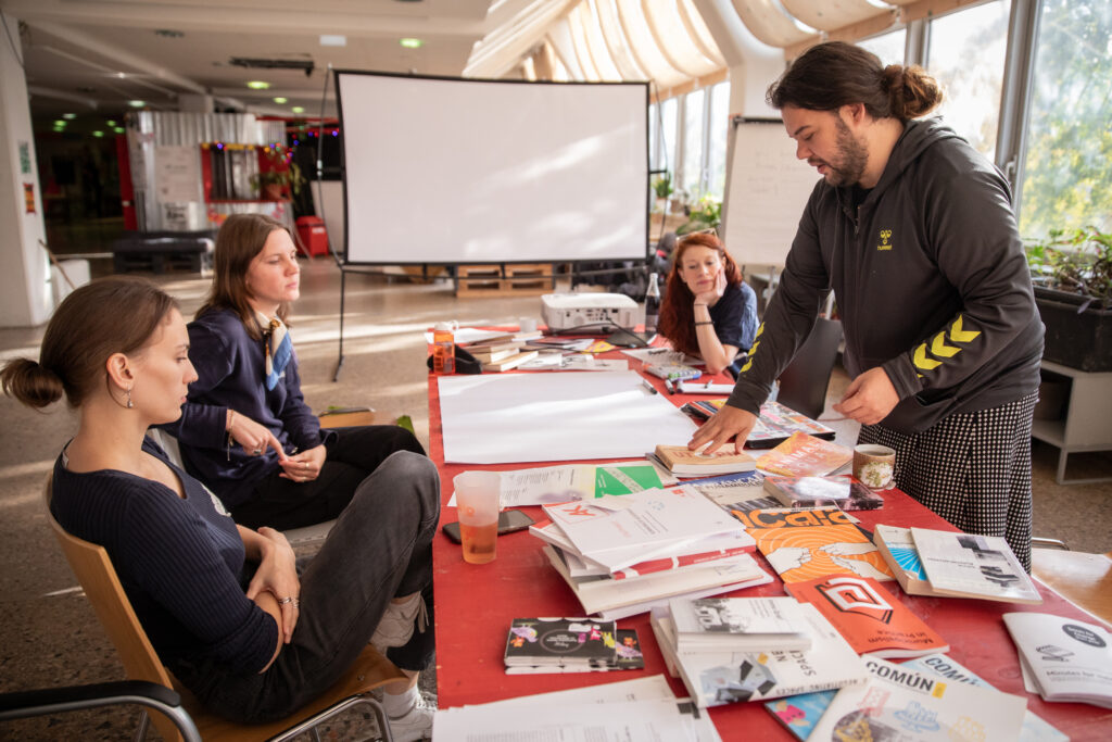 Eine Gruppe Menschen an einem Tisch die sich unterhält, im Hintergrund eine Leinwand. Auf dem Tisch diverse Materialien wie Bücher und Broschüren