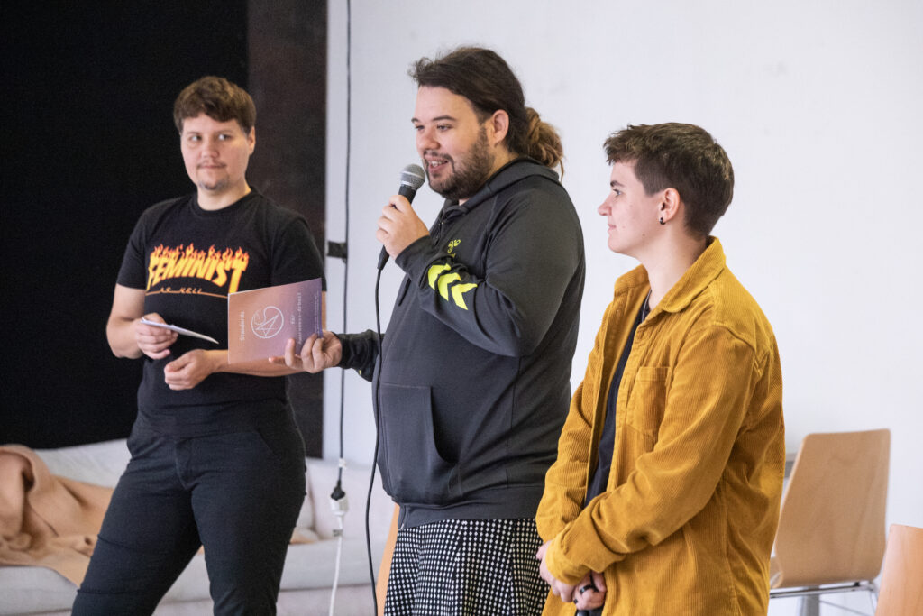 Three people are standing on a stage as speakers, with the person in the middle holding a microphone and speaking.